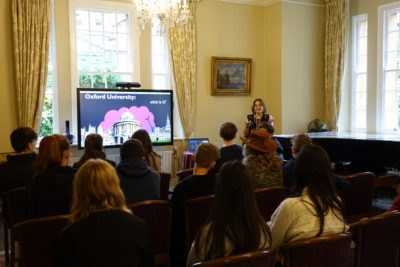 A student speaker stands at the front of a room presenting to a seated audience of prospective students. A large screen beside the speaker displays a slide titled ‘Oxford University: what is it?’ The room has tall windows with curtains, a chandelier, and a grand piano.