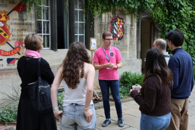 A student ambassador in a bright pink Exeter College T-shirt stands in front of an ivy-covered stone wall with painted college crests, talking to a small group of visitors. The visitors, a mix of adults and young people, listen attentively in an outdoor courtyard setting.
