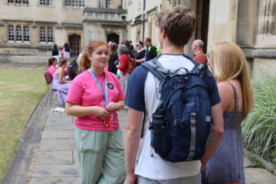 A student ambassador wearing a bright pink Exeter College T-shirt talks to two prospective students standing with their backs to the camera. They are outdoors on a stone path next to a college building covered with greenery. Other visitors and ambassadors are visible further along the path.
