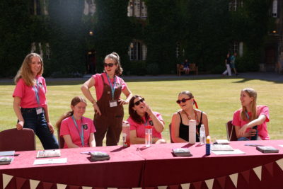 Six smiling student ambassadors wearing bright pink Exeter College T-shirts stand and sit behind a red table outdoors on a sunny day. They are in a college courtyard with green grass and ivy-covered stone buildings in the background. The table is set with stacks of prospectuses and informational flyers.