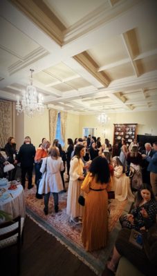 A crowded, elegant reception room where guests mingle under chandeliers, chatting and enjoying refreshments.