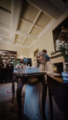 Melissa Thiesen sits at an ornate table signing copies of her book while guests gather around in a formal, historic room.
