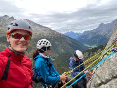 Three climbers wearing helmets and outdoor gear are roped together on a mountain cliff, securing their lines. The valley below and rugged peaks stretch into the distance under a cloudy sky.