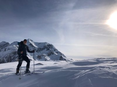 A skier stands on a snowy mountain slope with ski poles, surrounded by high alpine peaks under bright sunlight. The snow and light create a crisp, clear winter scene.