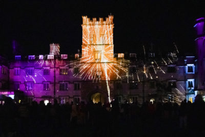 A night-time crowd watches vibrant light projections burst across the front of Exeter College’s tower and buildings, creating firework-like patterns in orange, purple, and blue.