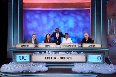The full Exeter College alumni team on the University Challenge Christmas special. Four team members sit behind labelled desks: “Watson,” “Berg,” “Chakrabarti,” and “Anson,” with the host standing behind the middle two. The set is decorated with winter and snowflake motifs, and the front panel reads “Exeter – Oxford".