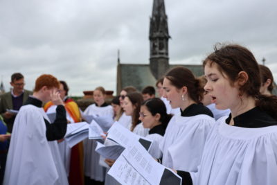 A group of choir singers dressed in white robes standing outdoors, singing from sheet music. The background features a church building with a tall spire under a cloudy sky.