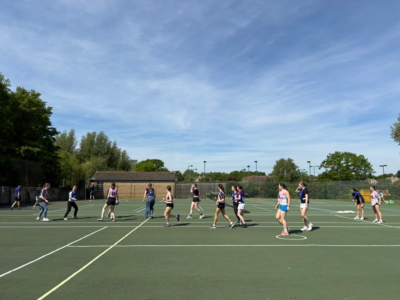A large group of people playing a team sport on an outdoor netball court under a bright blue sky, with players spread across the court and trees in the background.