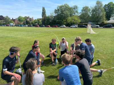 A group of people kneeling in a circle on a grassy sports field holding lacrosse sticks, appearing to discuss strategy or take a team break.