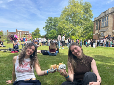 Two young women sitting on grass at an outdoor campus-style event, smiling while holding burgers, with crowds of people and historic-looking buildings behind them.