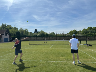 People playing doubles tennis on a sunny grass court, with one player serving the ball while others wait in position under a clear sky.