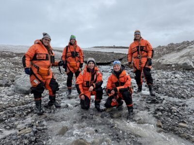 Five members of the Svalbard expedition, including Dr Lukas Krone, wearing bright orange cold-weather suits, pose together in a rocky glacial stream surrounded by snow-covered terrain