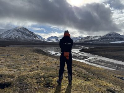 Dr Lukas Krone seen from behind wearing a jacket marked “EXETER”, looks across a broad Arctic valley towards snow-covered mountains and a winding glacial river.